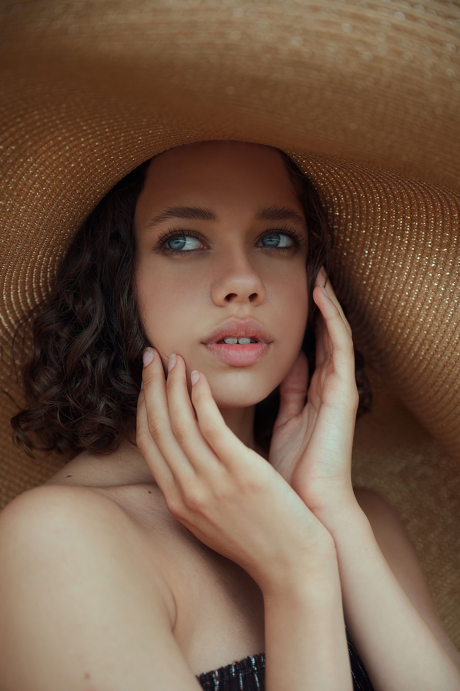 Romantic portrait under wide-brim straw hat with soft lighting and natural makeup.