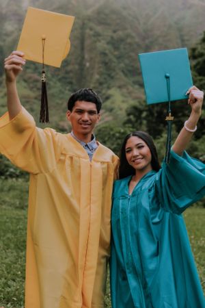 Graduates in yellow and teal gowns holding up their diplomas against a mountain backdrop.