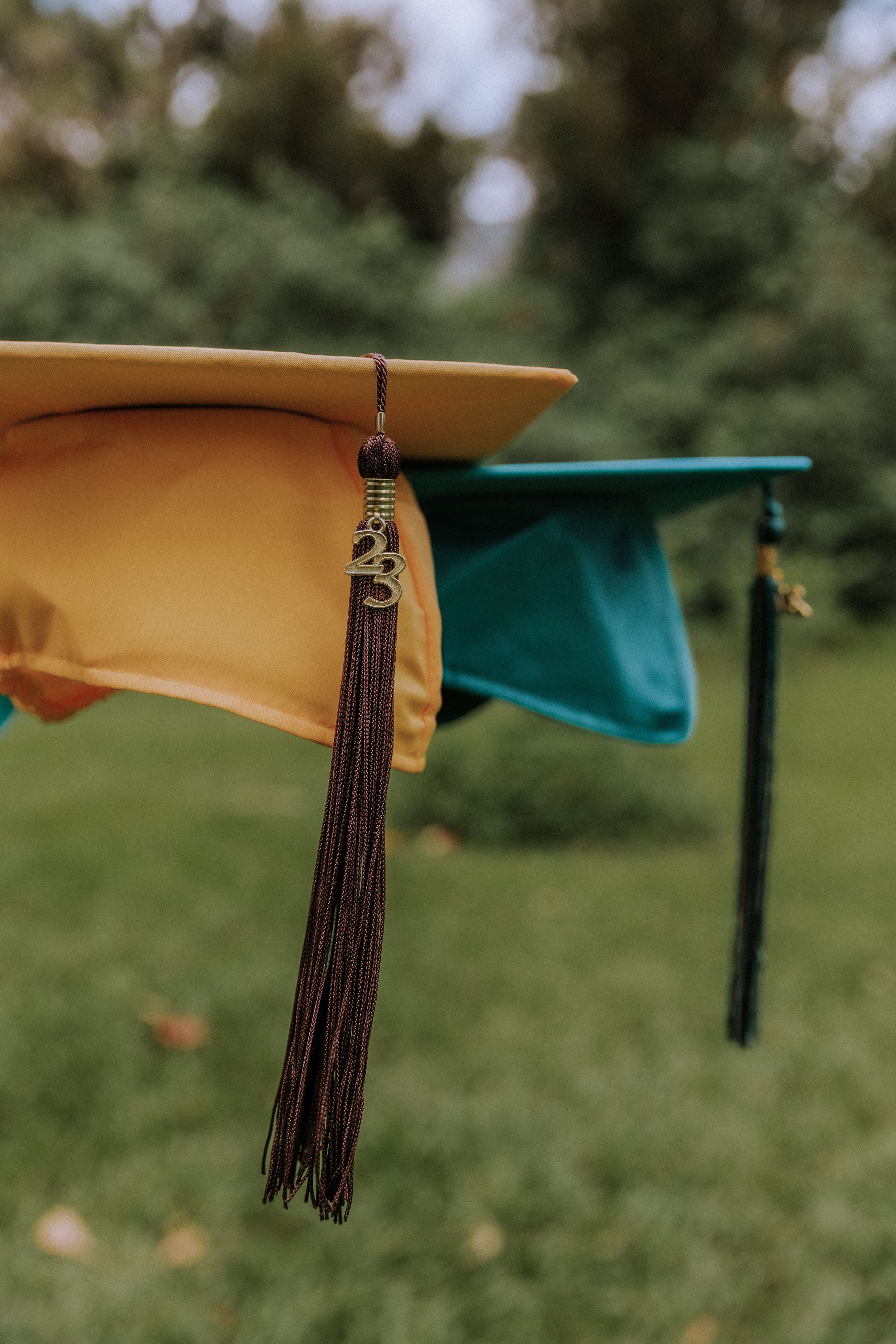 Close-up of graduation cap tassels in gold and teal colors against a blurred natural background.
