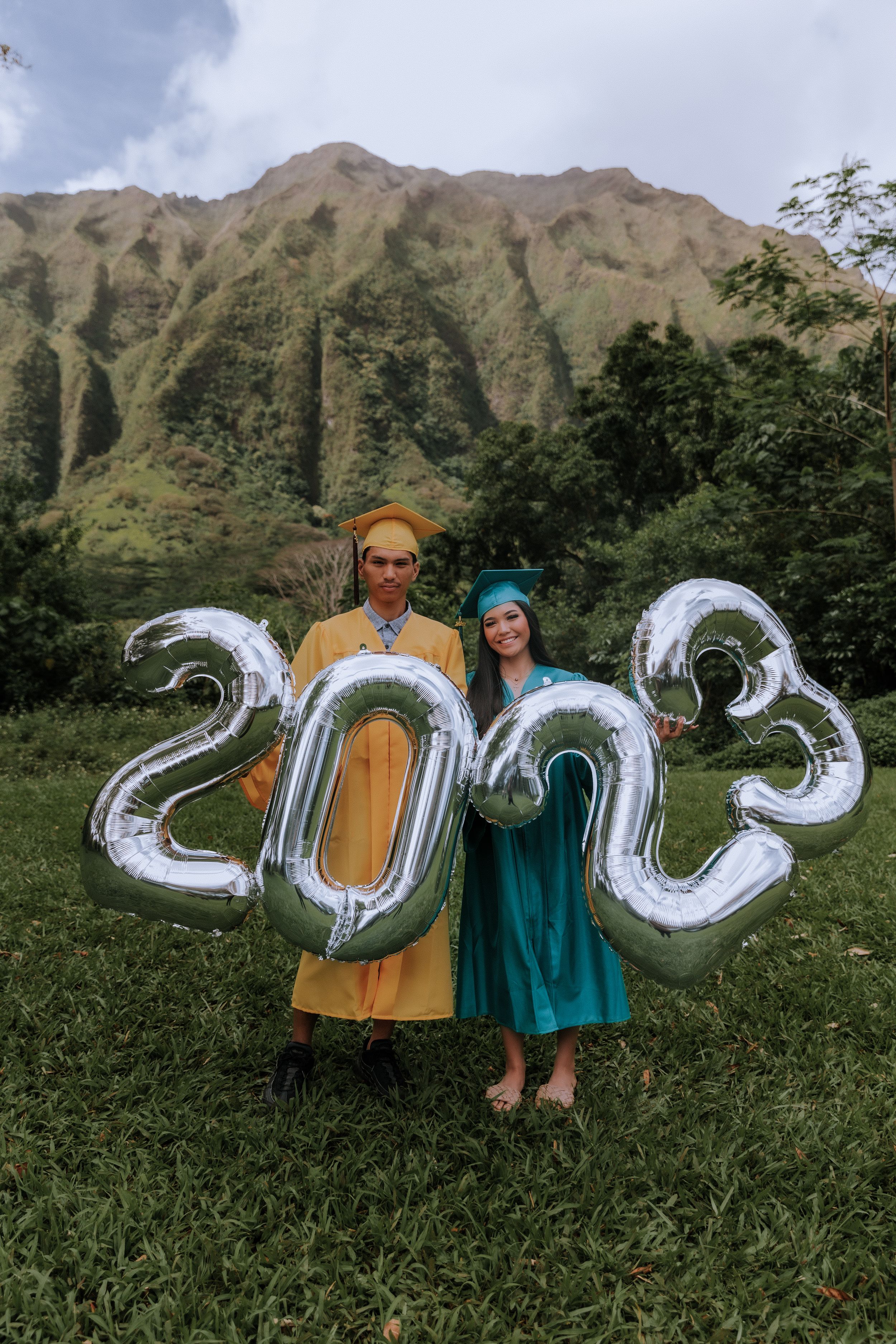 Two people in graduation attire hold silver 2023 balloons against a mountainous backdrop.