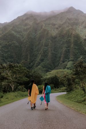 Two graduates walking down a scenic mountain road carrying colorful graduation gowns.