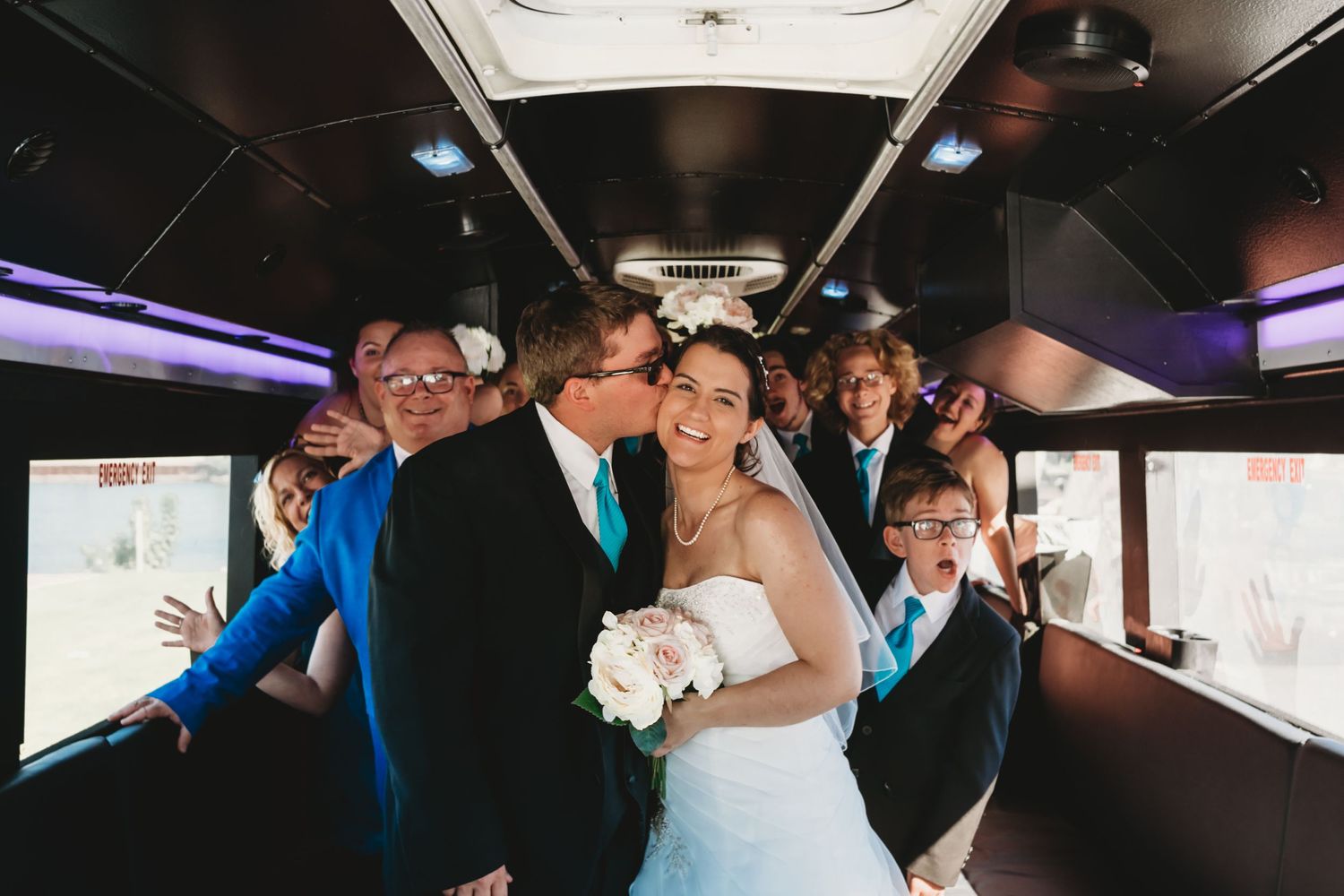 A wedding party celebrates inside a party bus with purple accent lighting.