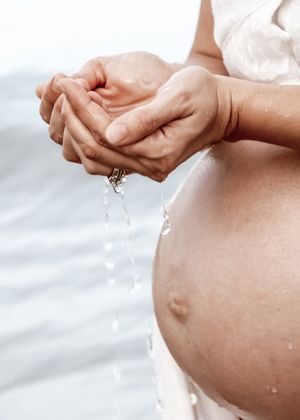 Hands cupping water over pregnant belly with water droplets falling against white background.