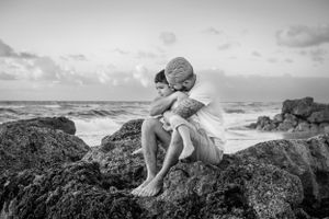 Black and white photo of figure sitting contemplatively on coastal rocks by ocean.