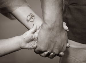 Close-up black and white photo of two hands clasped together with a small heart tattoo visible on one wrist.