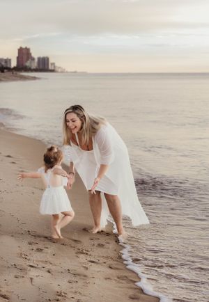 Mother and child playing together on beach at sunset wearing white dresses.