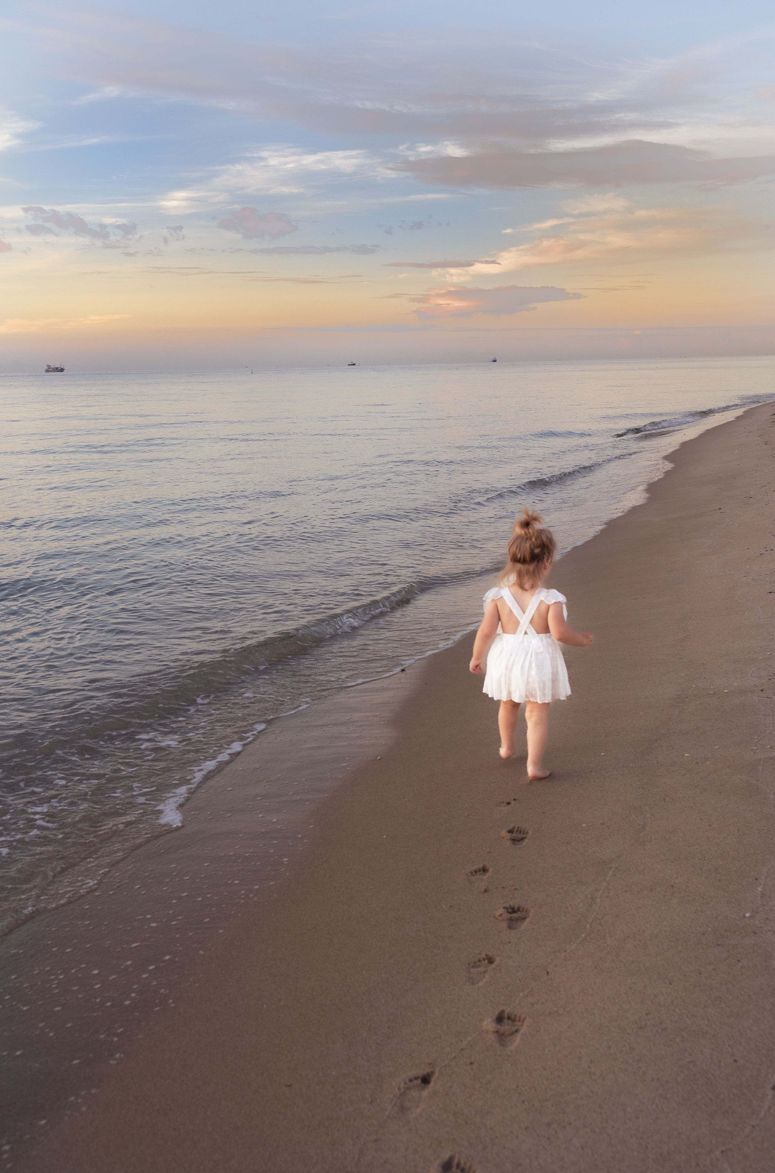 Small child in white dress leaves footprints while walking along empty beach at sunset.