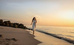 Ethereal figure in white dress walks along beach at sunset casting long shadows on golden sand.