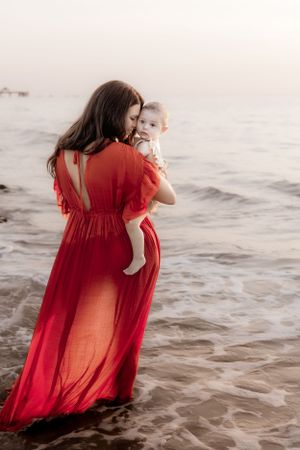 Figure in flowing red dress holds child while standing on beach at sunset.