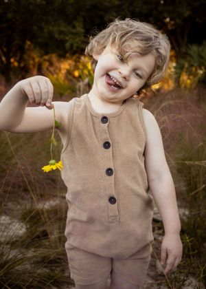 Child wearing tan button-up outfit laughing while holding yellow flower outdoors.