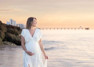 Expecting mother in white dress standing on beach at sunset with pier in background.