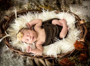 Sleeping newborn wrapped in brown swaddle resting in wicker basket on white fur.