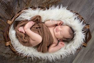 Sleeping newborn baby wrapped in brown fabric lying in white fur-lined wooden basket.