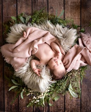 Newborn baby sleeping on white fur surrounded by greenery and pink blankets in rustic wooden setting.