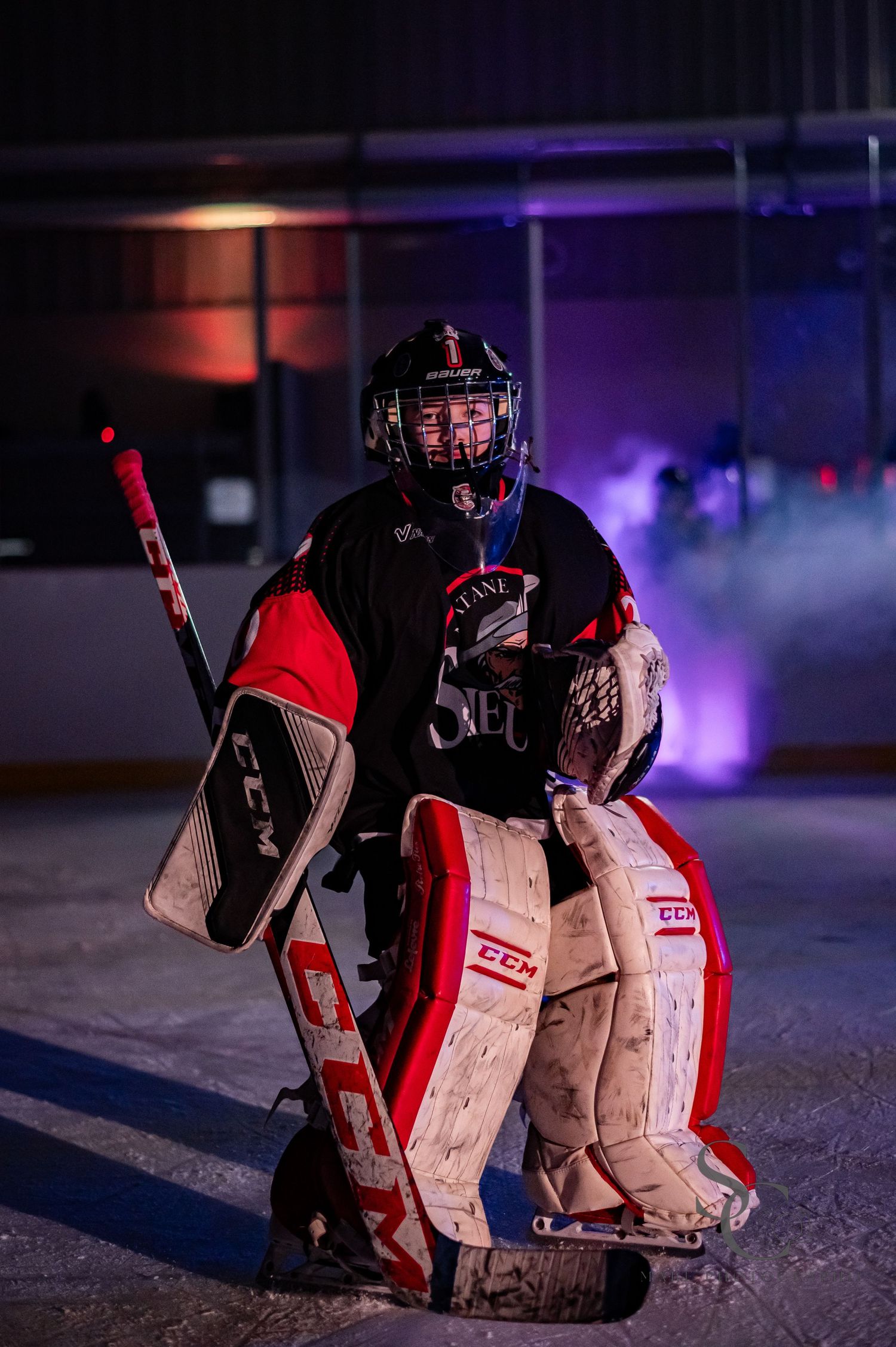 Ice hockey goalie in red and black gear crouching on ice with dramatic purple and red lighting.