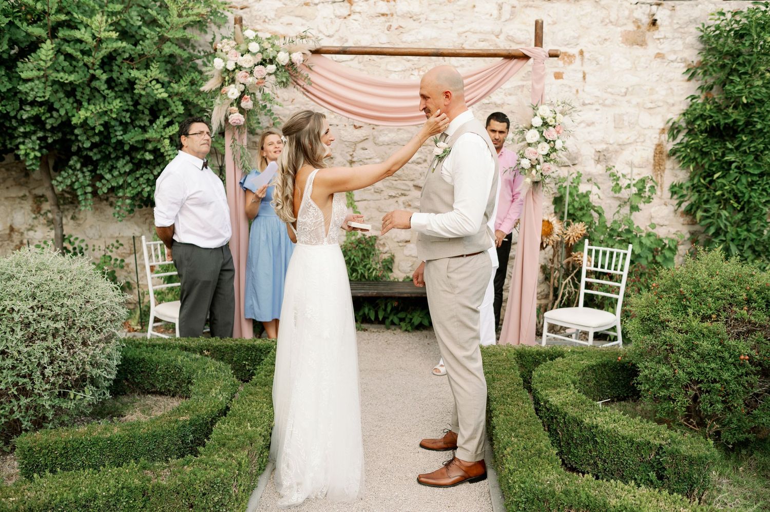 Couple shares emotional moment during garden wedding ceremony with manicured hedges and peachy backdrop.