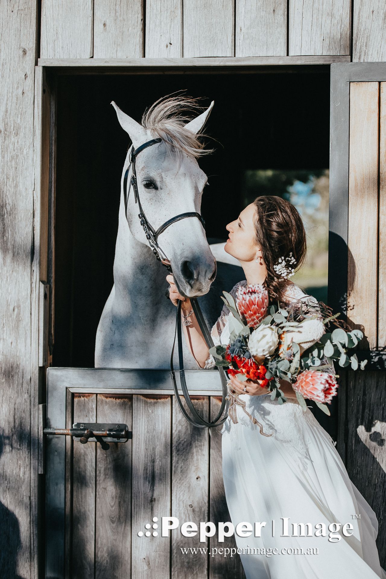 A bride in a white wedding dress kisses a white horse while holding a floral bouquet outside a rustic wooden stable.