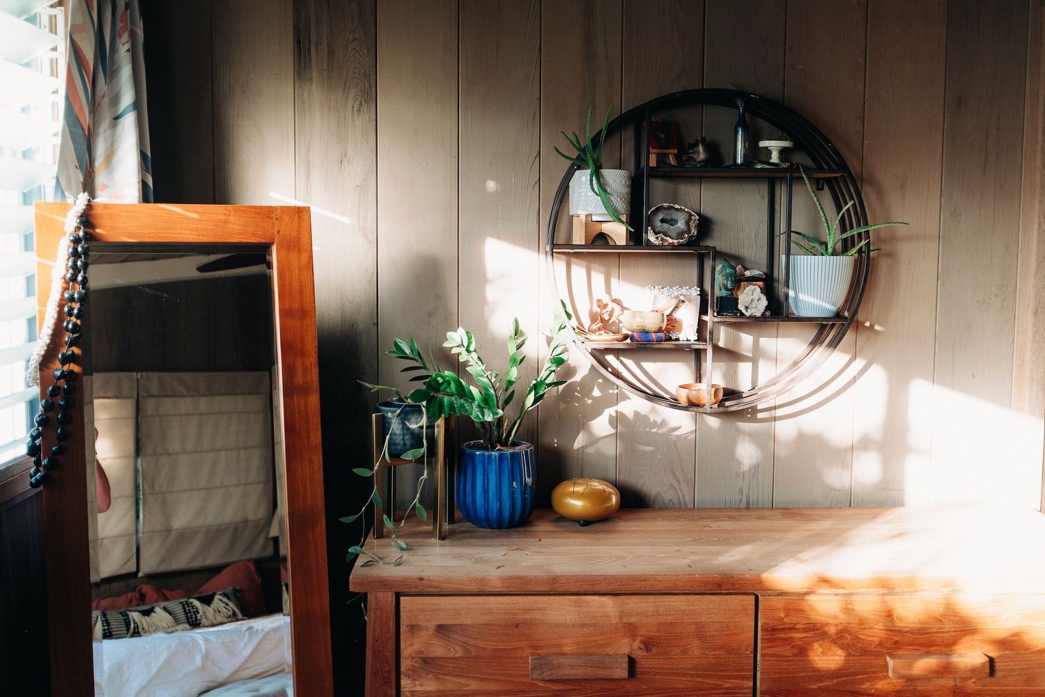 Sunlit interior with wooden dresser, round mirror, and decorative plants casting shadows.