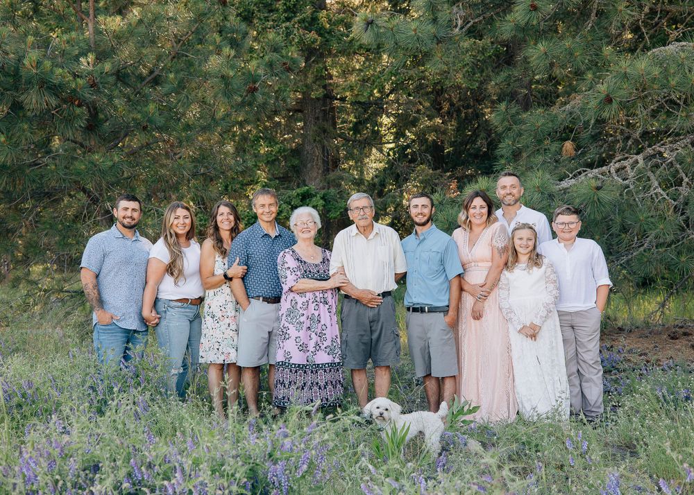 Extended family poses together outdoors in a natural setting with purple wildflowers and trees in the background.