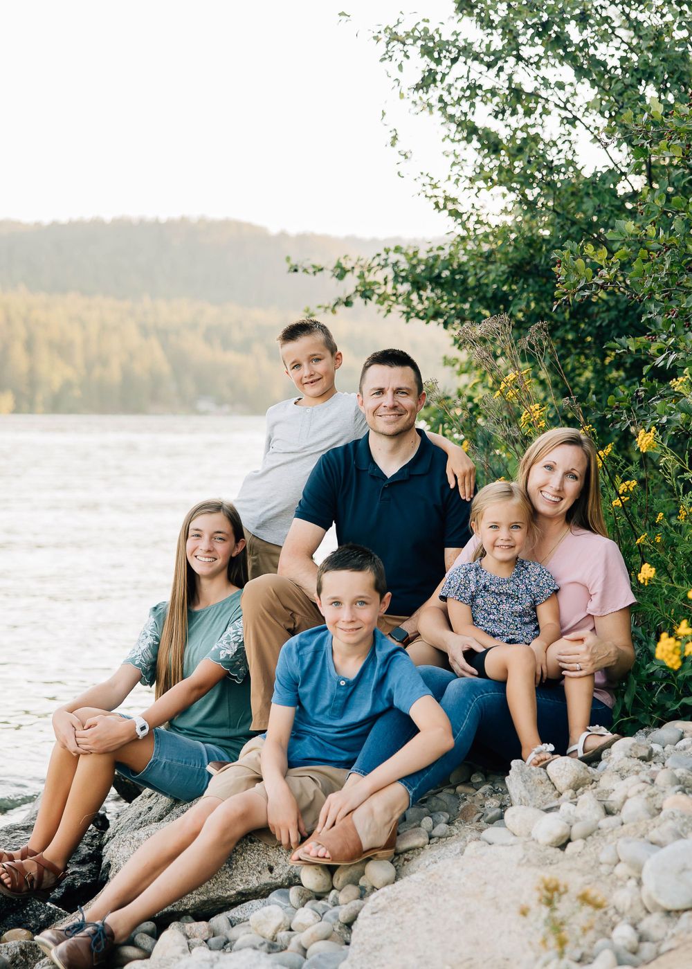 Family of six sitting together on rocky shore with river and forest in background.