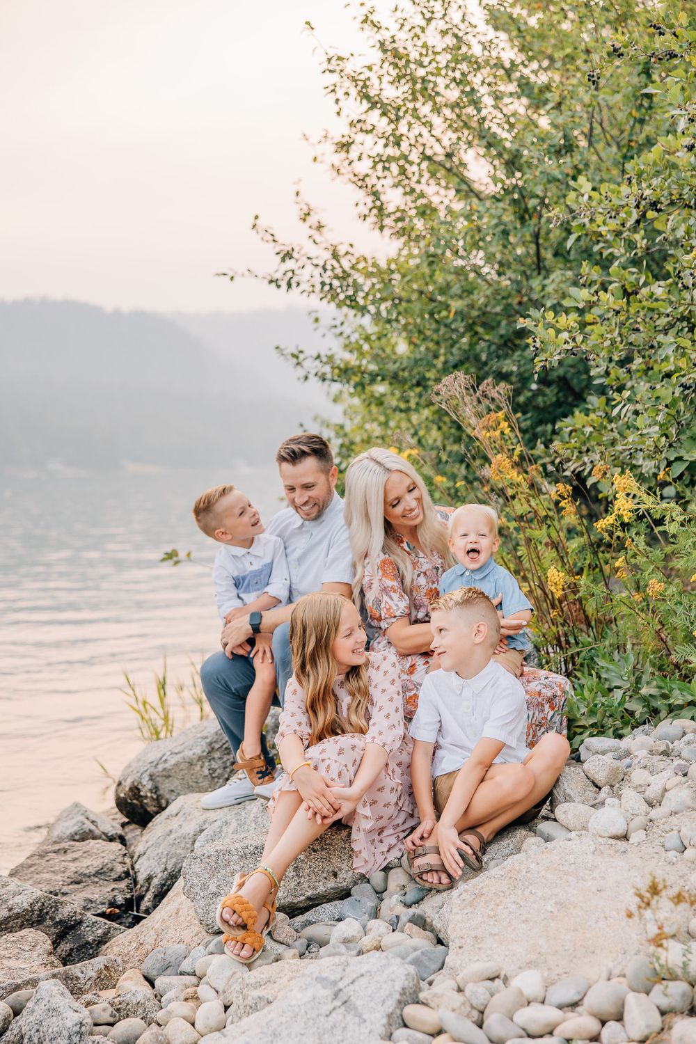 Family relaxing together on rocky shoreline during golden hour with lake in background.