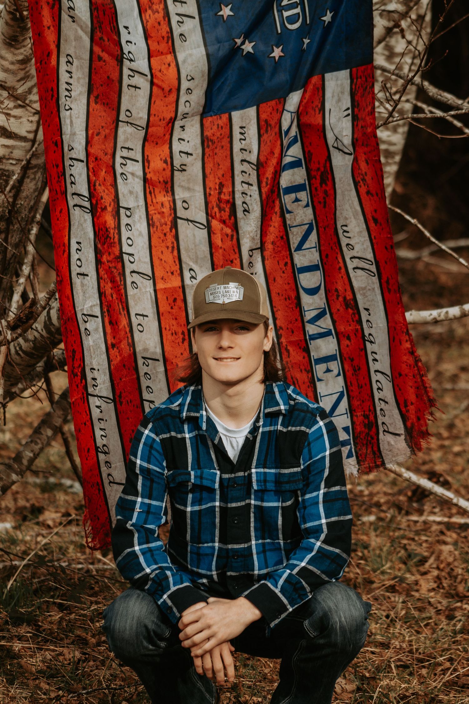 High School senior in a baseball cap and plaid flannel shirt poses in front of a rustic American flag decoration in an outdoor setting.