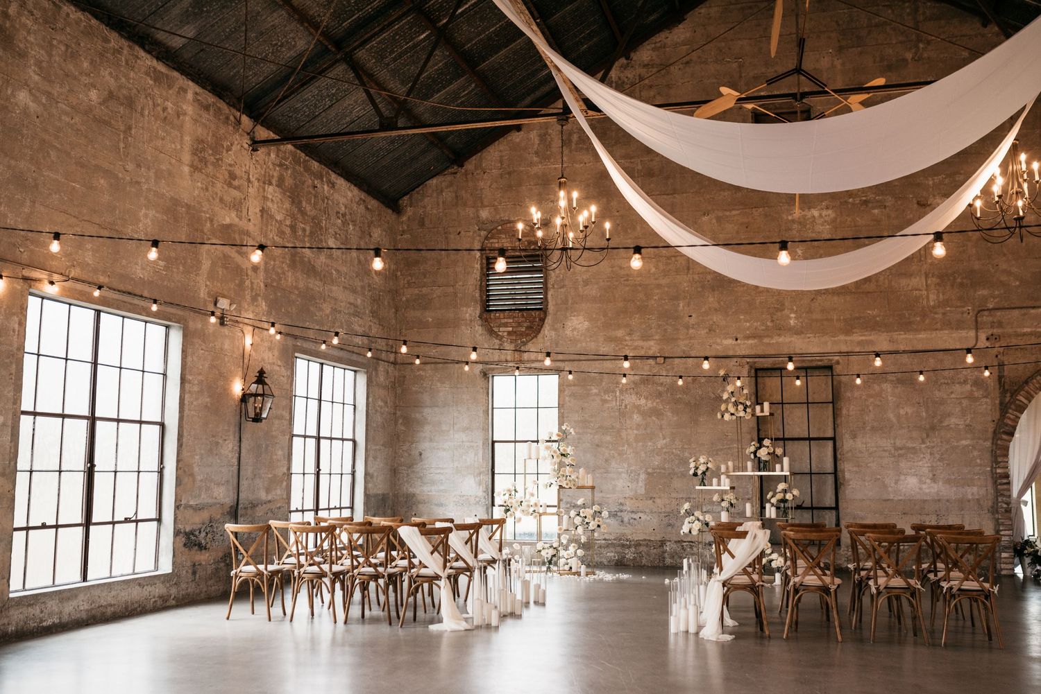 Industrial chic wedding venue with draping white fabric, string lights and wooden chairs.