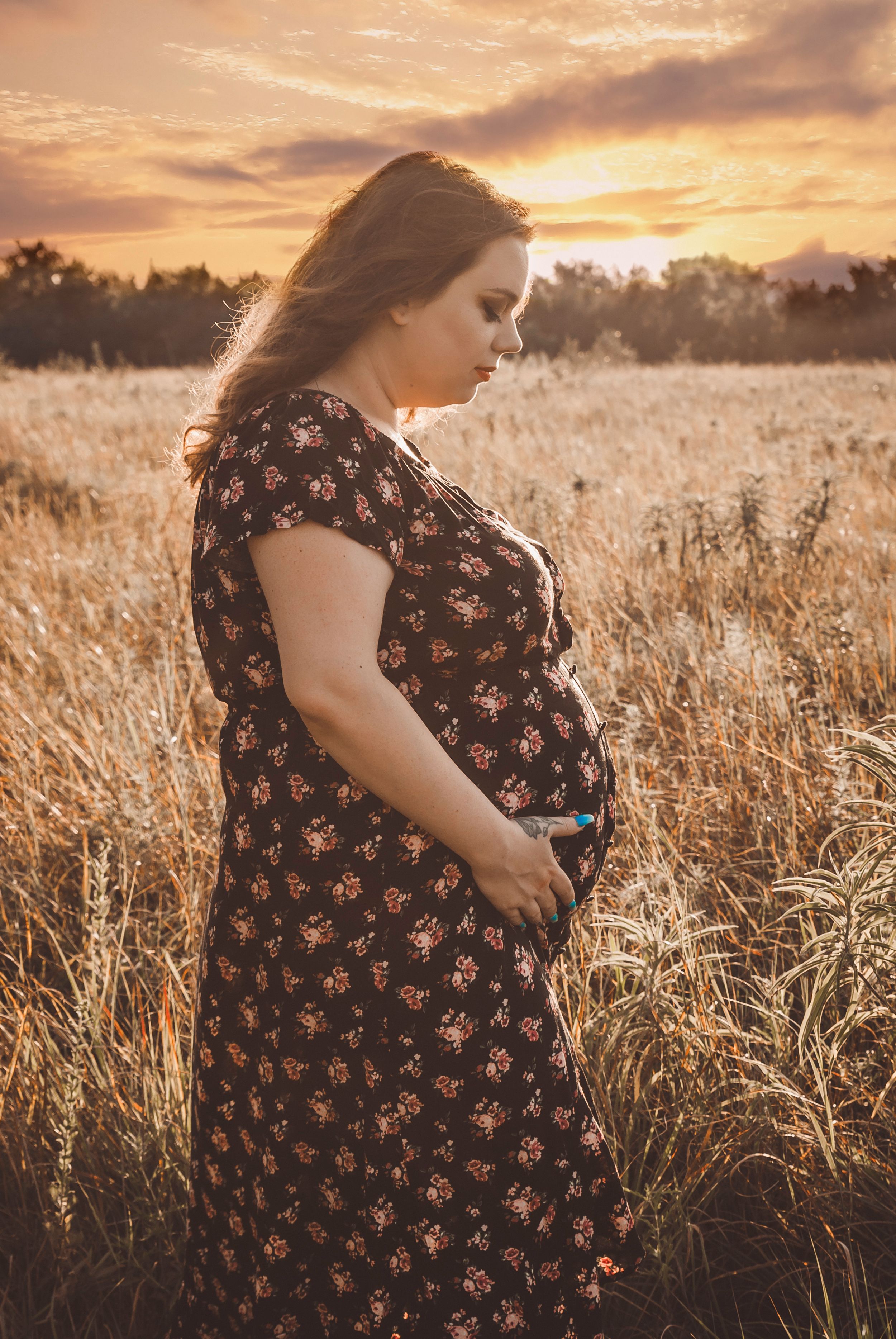 a maternity photo in an open field with a woman holding her belly and looking down at it lovingly with the sun coming in behind her