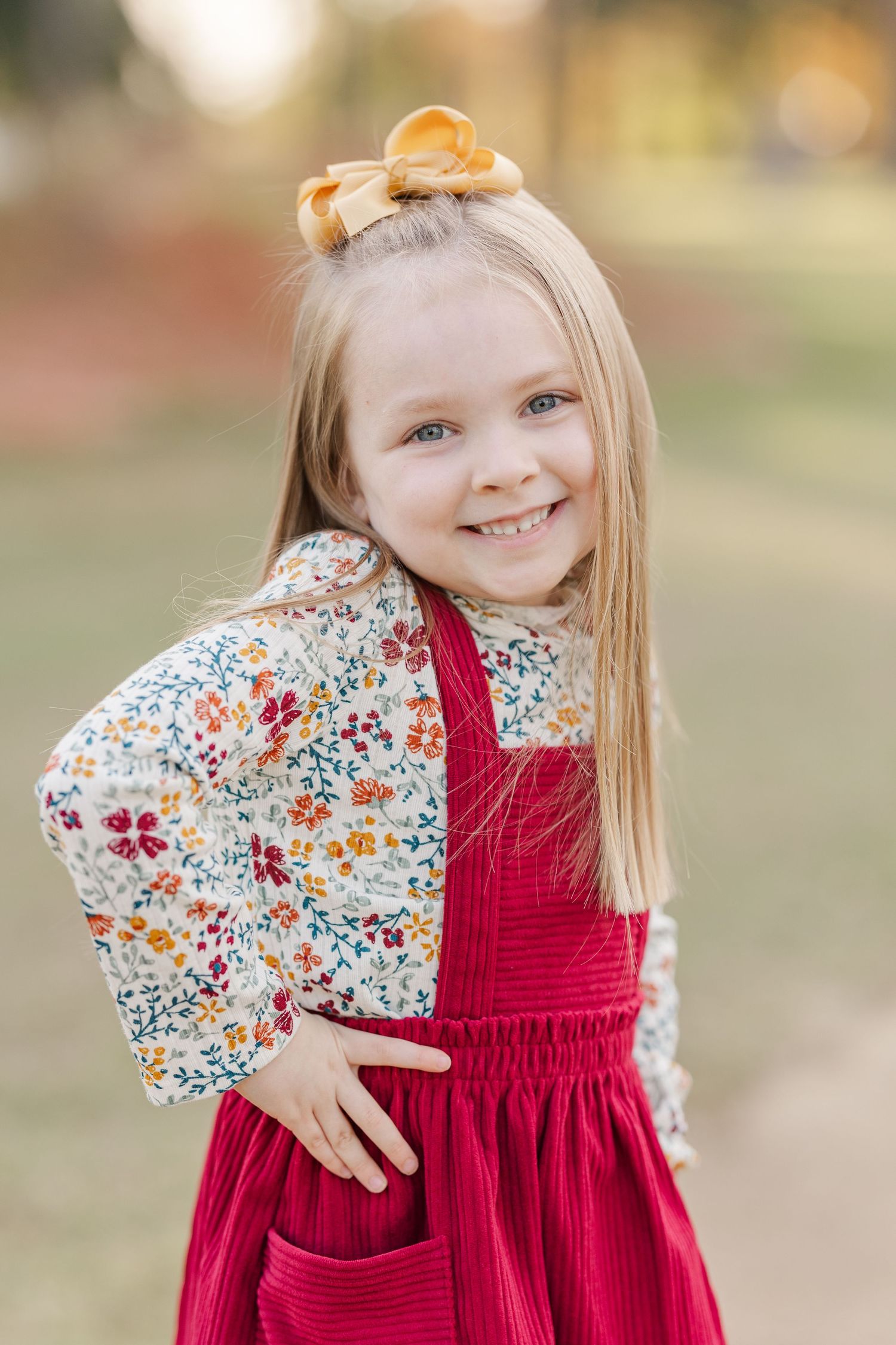 Young girl wearing floral top and red jumper dress with bow in hair posing outdoors.
