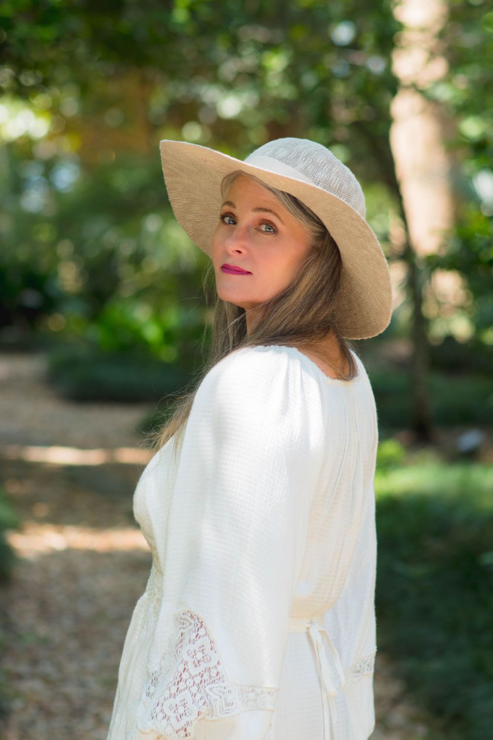 A woman in a white dress and wide-brimmed hat poses gracefully in a sunlit garden path.