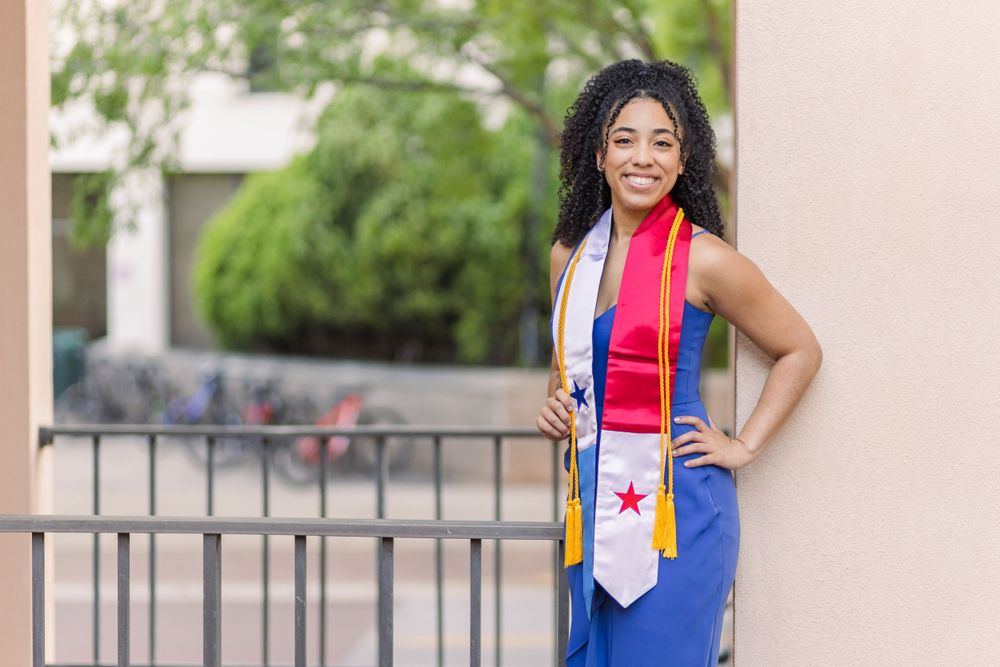 Graduate wearing a blue dress and graduation stole poses confidently outdoors.