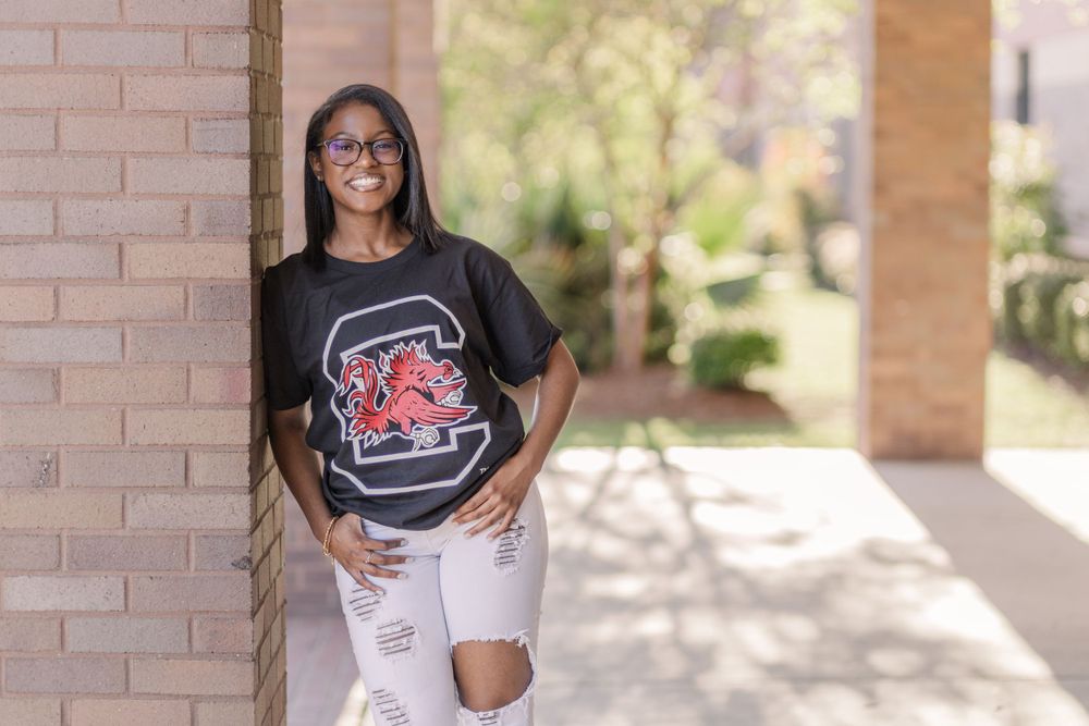 Student in university logo t-shirt and ripped jeans poses by brick wall.