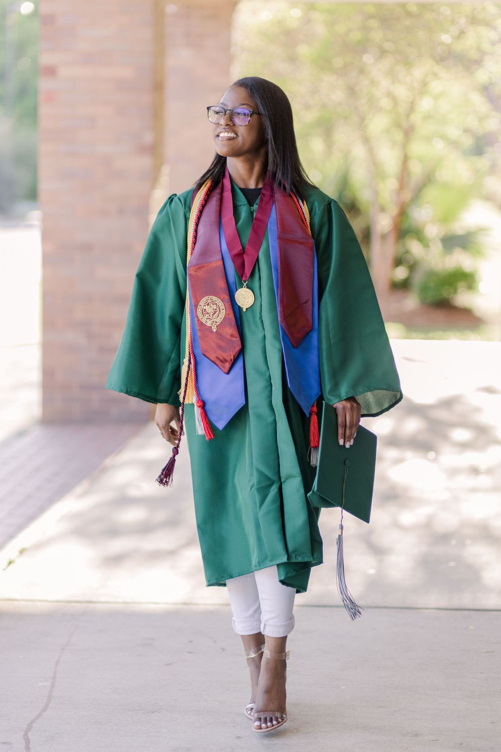 Graduate walks confidently in green regalia adorned with colorful stoles and medals.