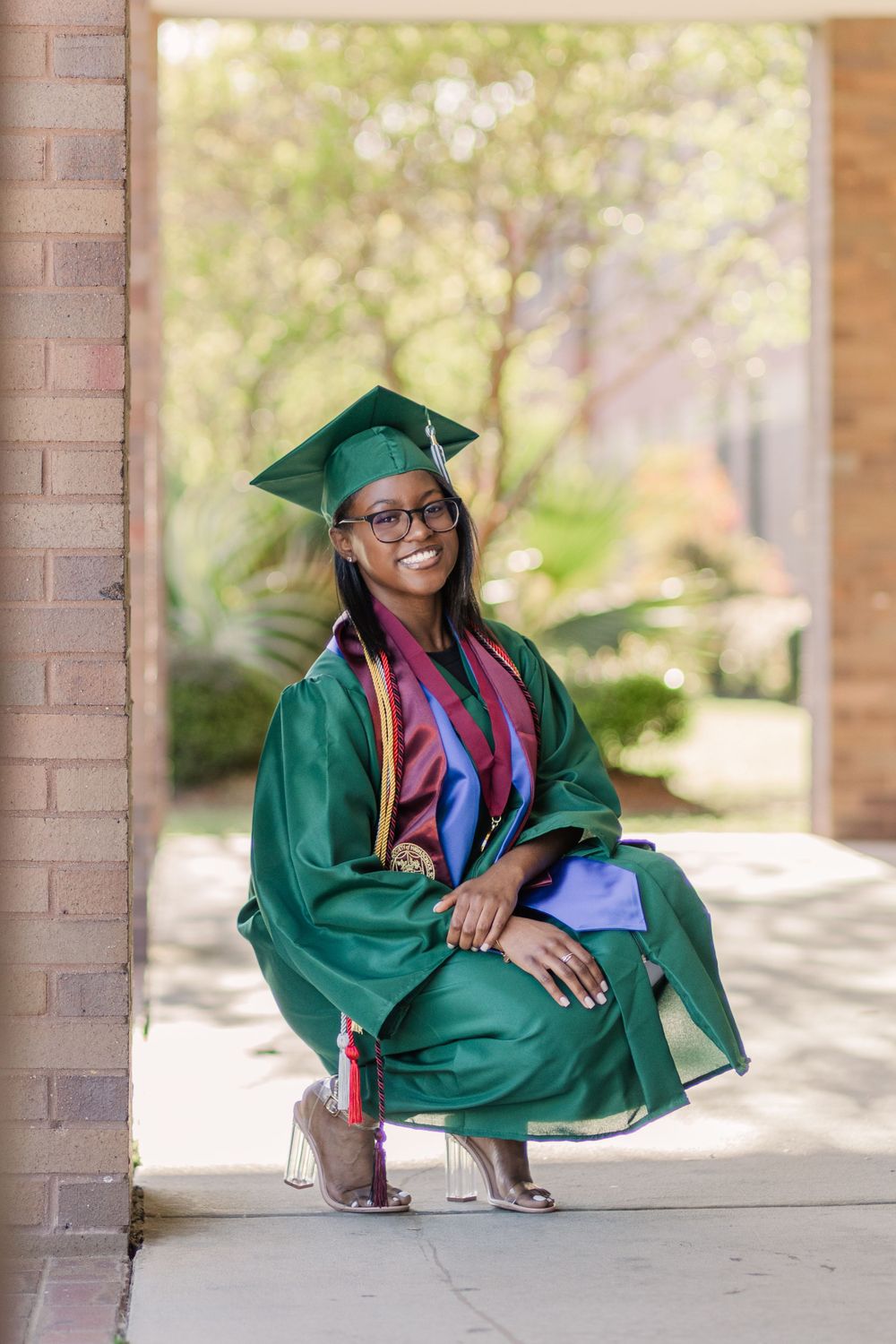 Graduate in emerald green cap and gown poses elegantly under archway.