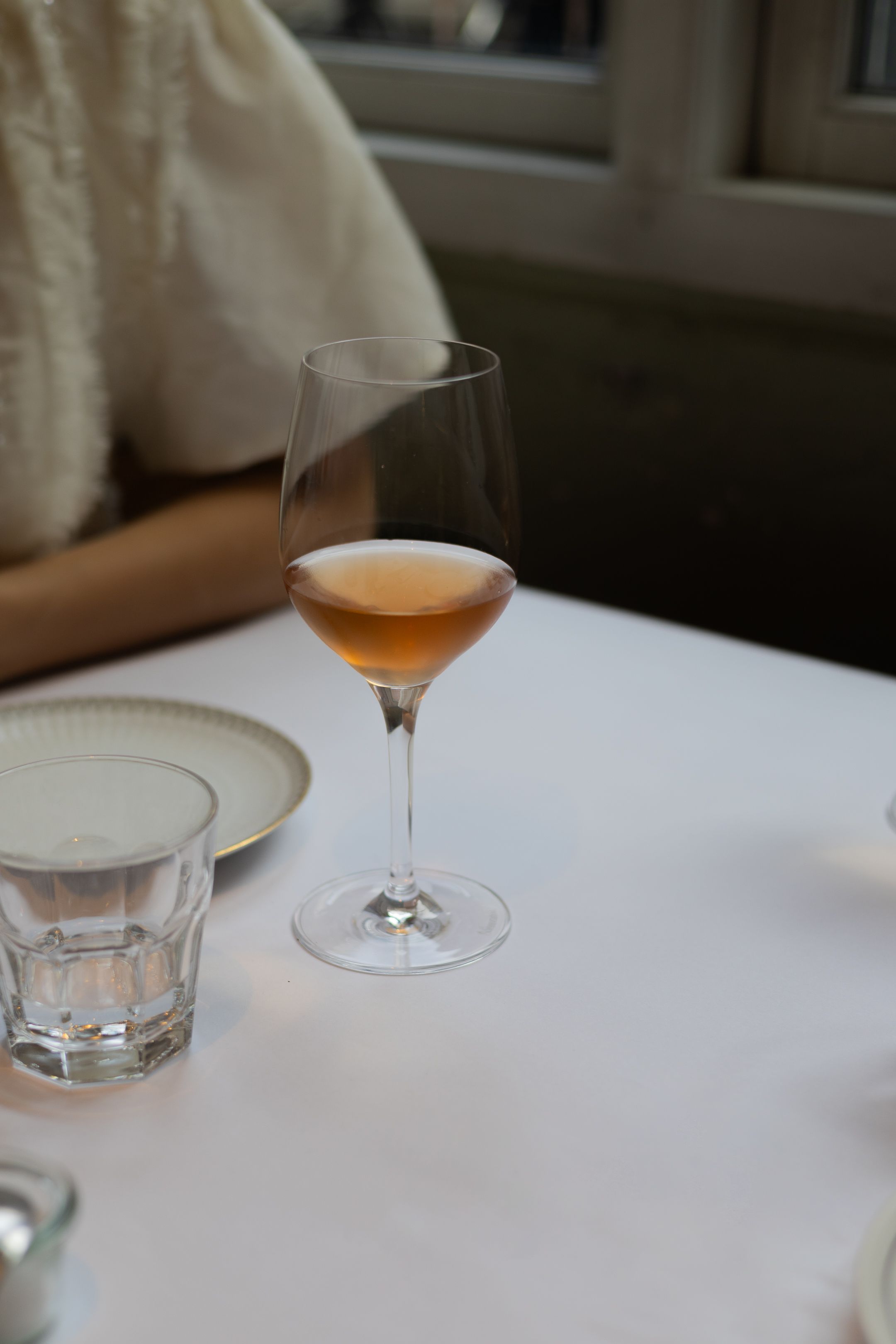 A glass of orange-tinted wine and a water glass sit on a white tablecloth in a restaurant setting.