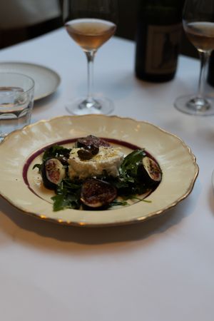 A plated dish of grilled figs and greens on a cream-colored plate with wine glasses in the background.
