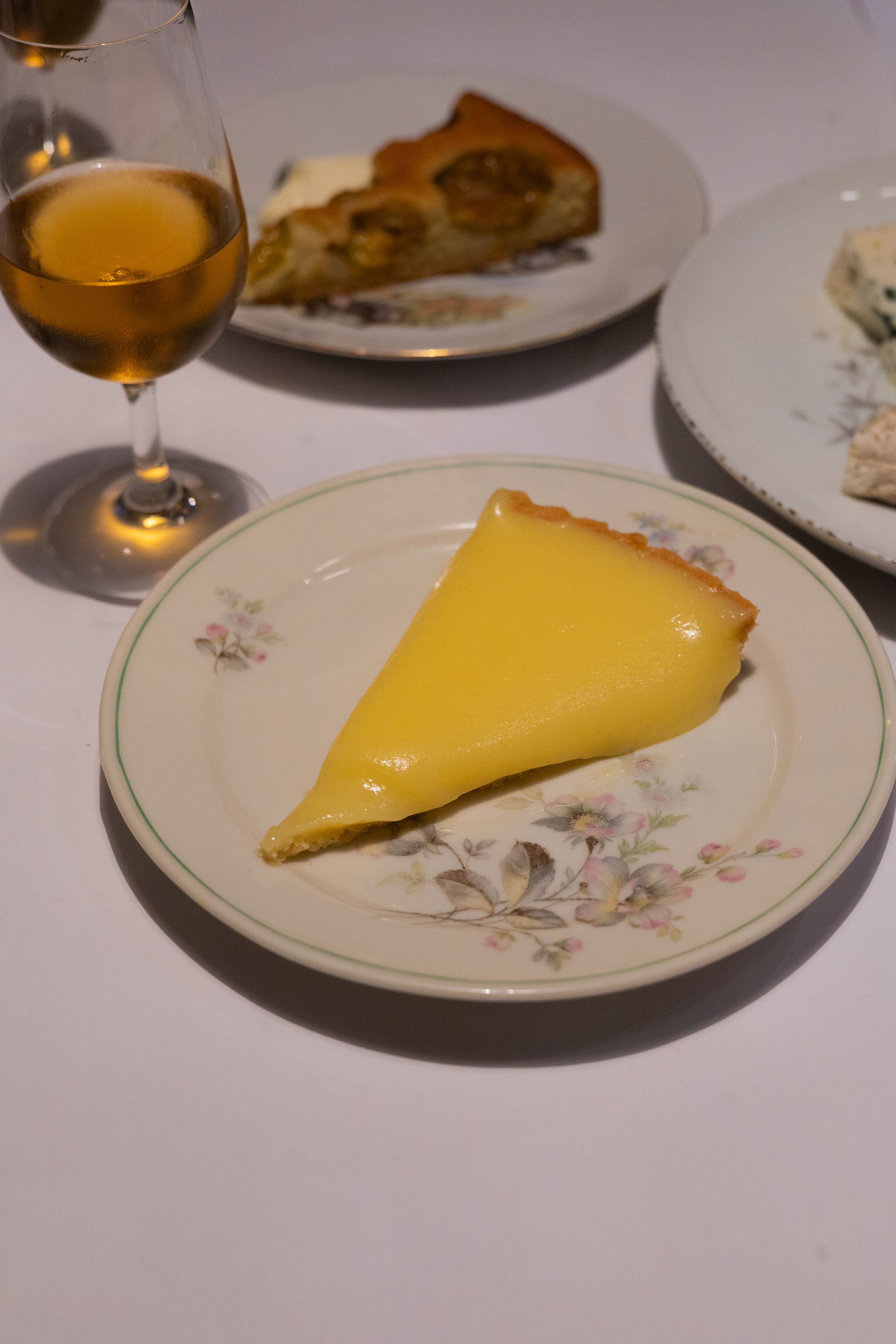 Slice of lemon tart on floral plate with glass of white wine.