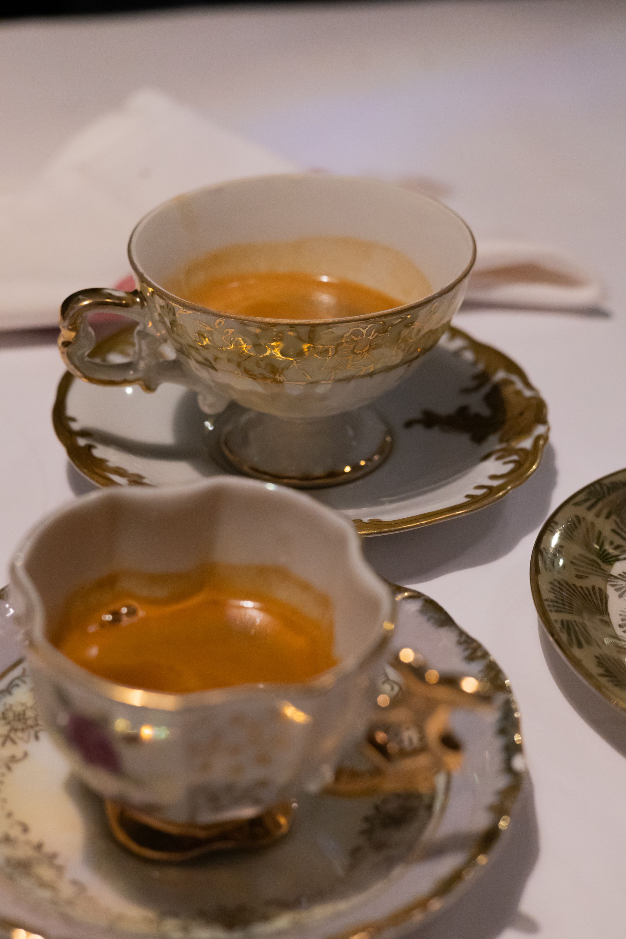 Two ornate vintage teacups with gold trim containing amber-colored tea on matching saucers.