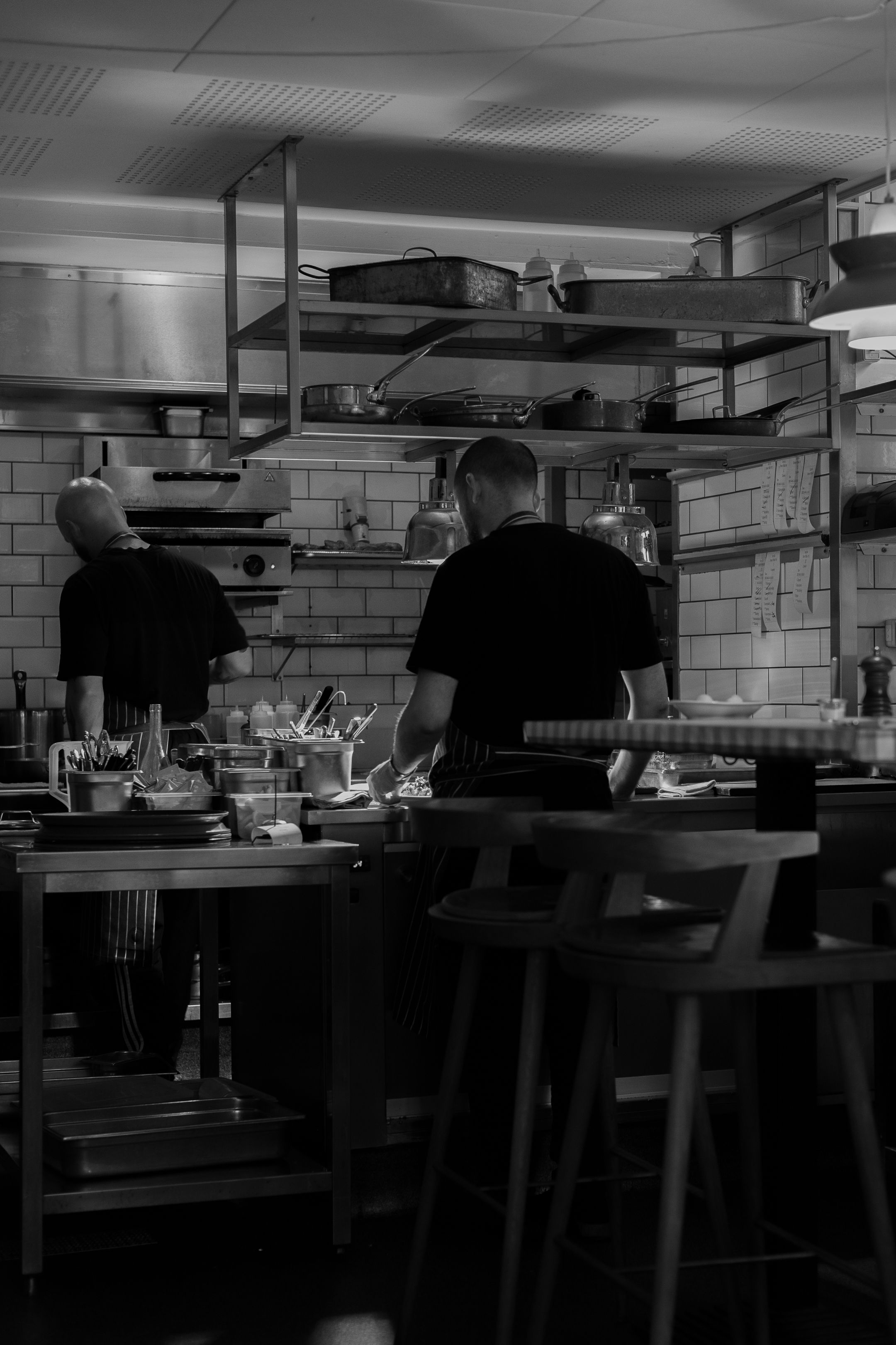 Black and white photo of busy restaurant kitchen with industrial shelving and staff at work.