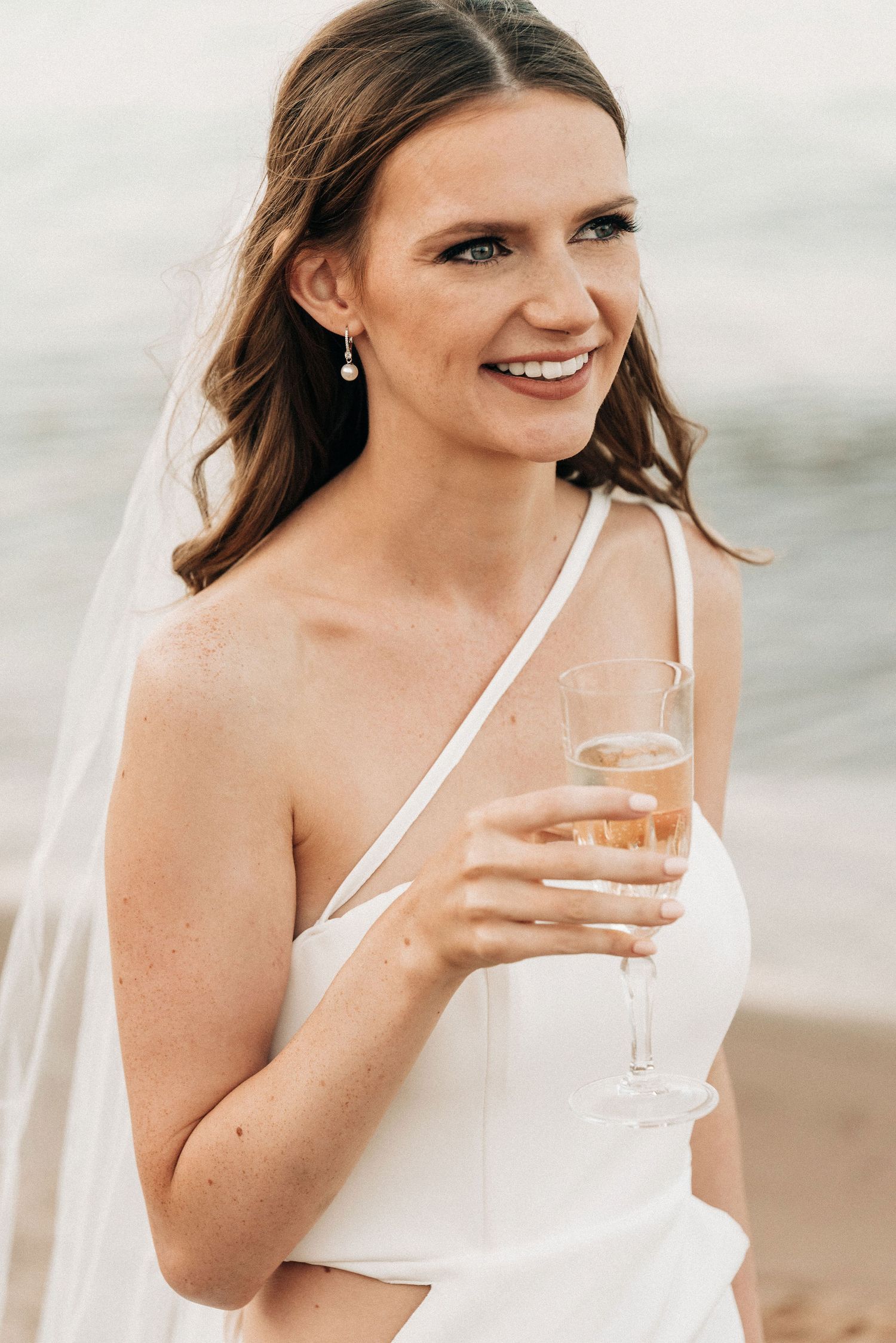 Bride in elegant white dress holding champagne glass by the ocean.