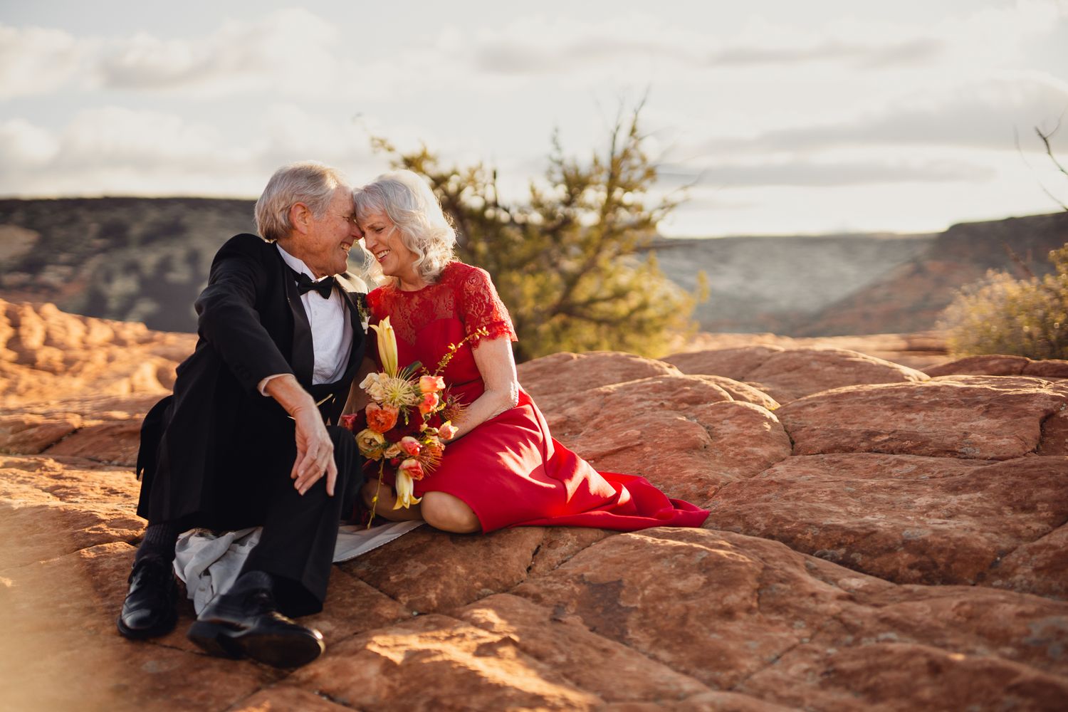 Elderly couple in formal wear share a romantic moment on red rock formations at sunset in the desert.