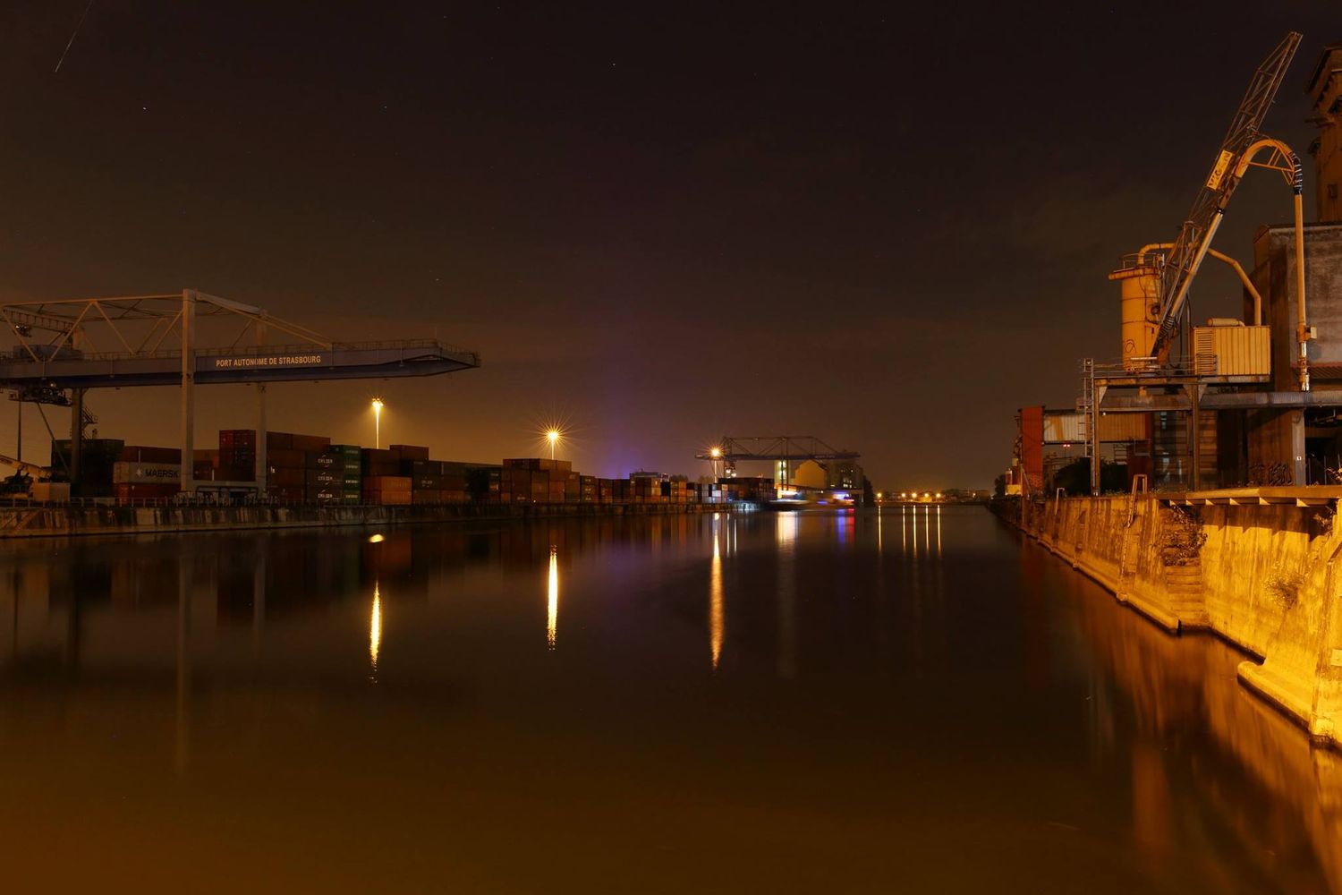 Industrial port at night with illuminated cranes and bridges reflecting on calm water.