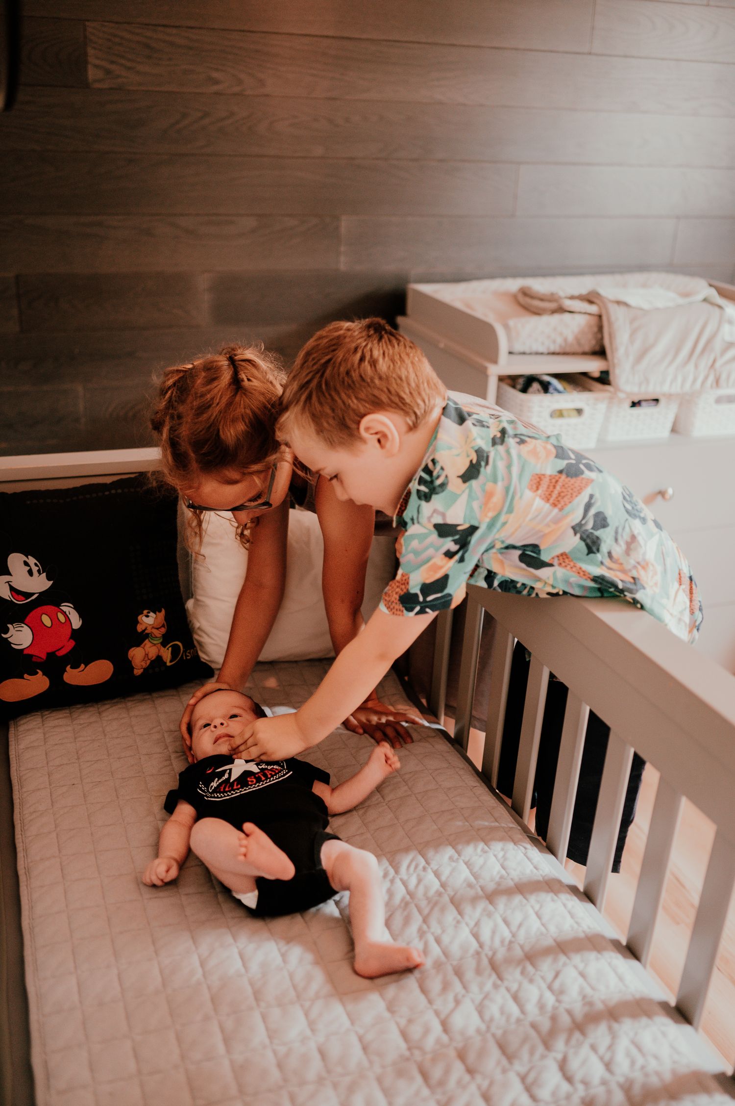 Parent reaches down to play with baby lying in white crib with Mickey Mouse plush toy nearby.