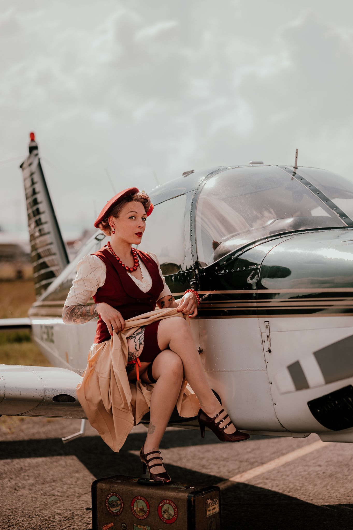 Vintage-styled woman in red beret poses on suitcase next to small private airplane.