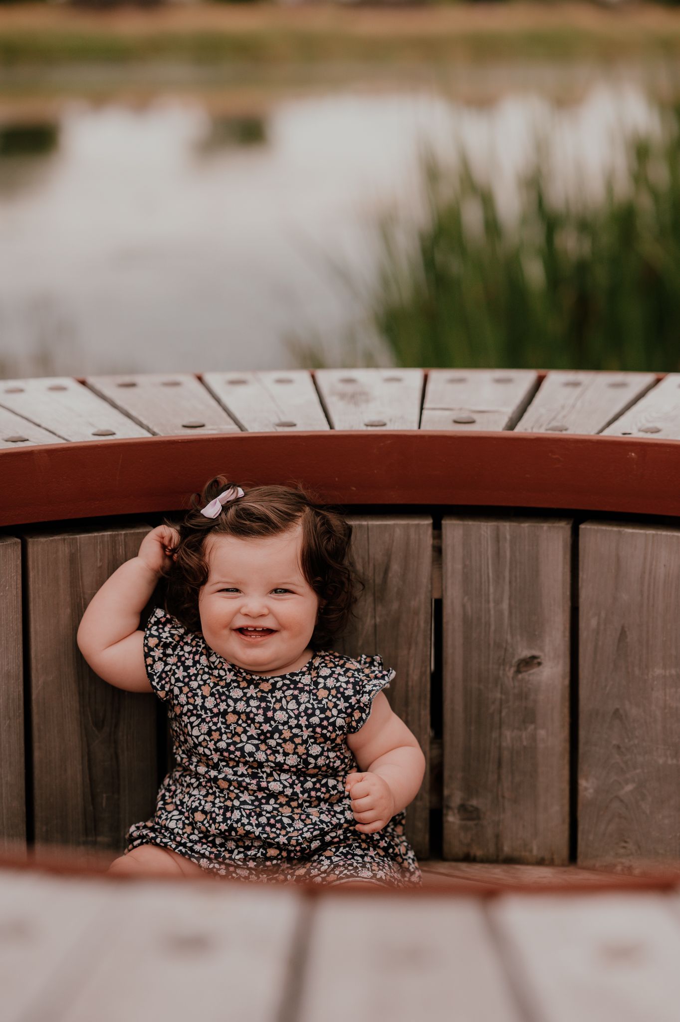 Smiling toddler in floral dress peeks over wooden railing by a pond.