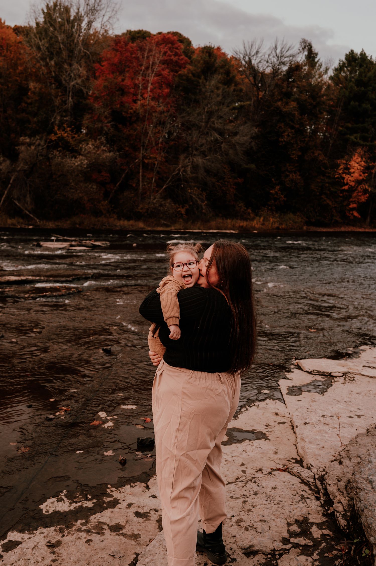Mother and child share tender moment by autumn riverbank with colorful foliage.