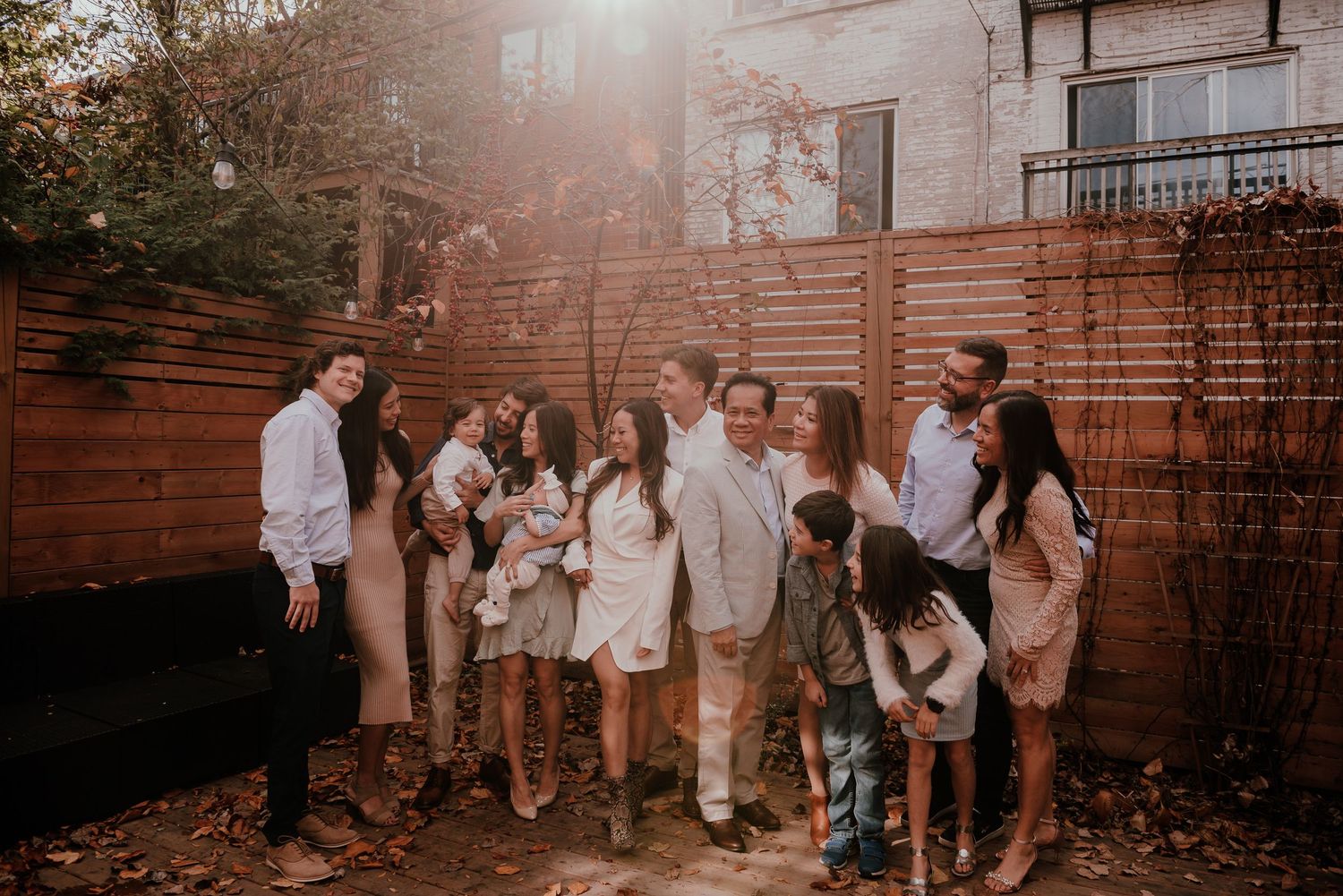 A warm sunlit gathering of people in casual white outfits standing together in a backyard with wooden fencing.