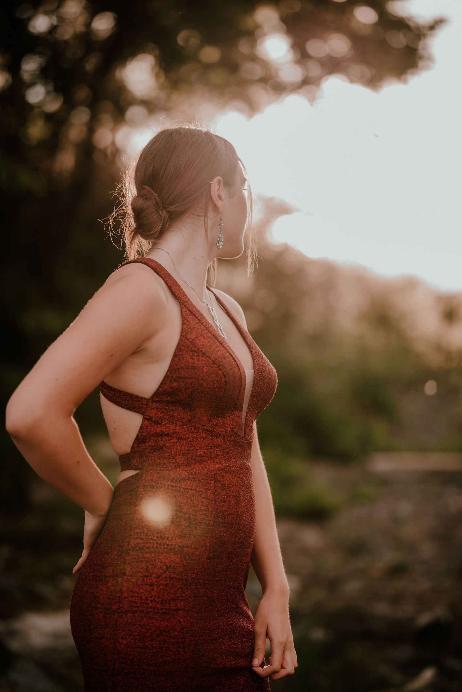 A woman in a burgundy dress poses at sunset with warm golden bokeh in the background.