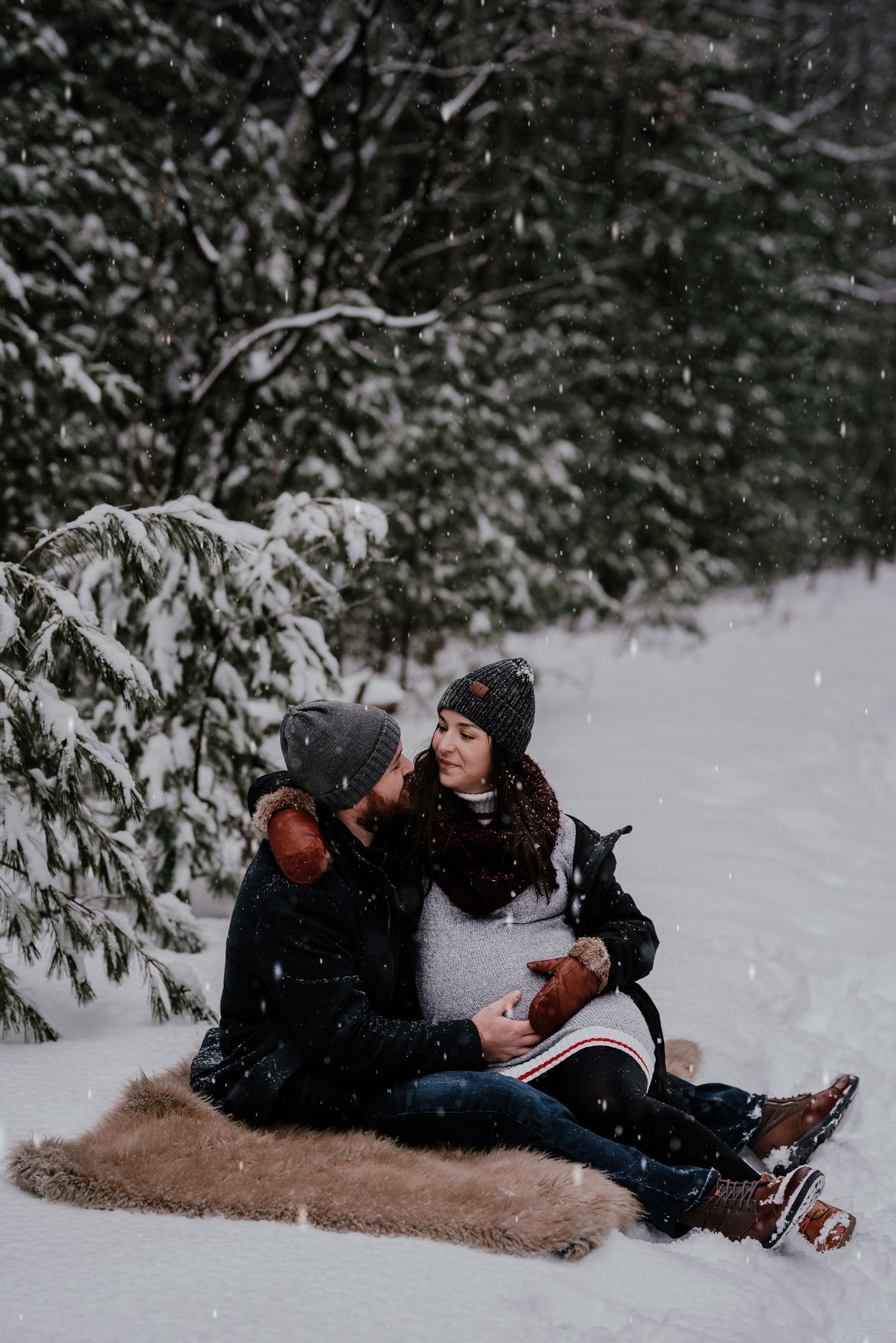 A couple sits together on a blanket in a snowy forest setting.