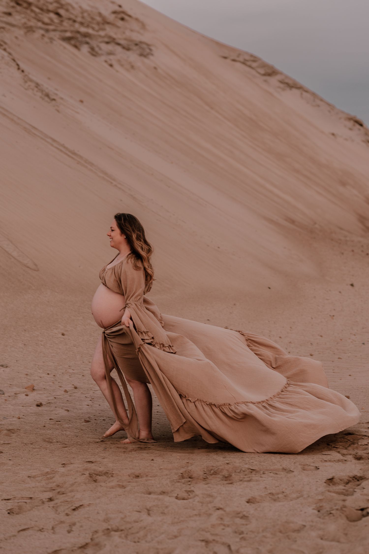 A maternity photo featuring a flowing beige dress against sand dunes at sunset.
